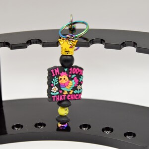 May include: A black beaded keychain with a gold crown charm and a pink, yellow, and blue charm with the text "I'm 100% That Chick".