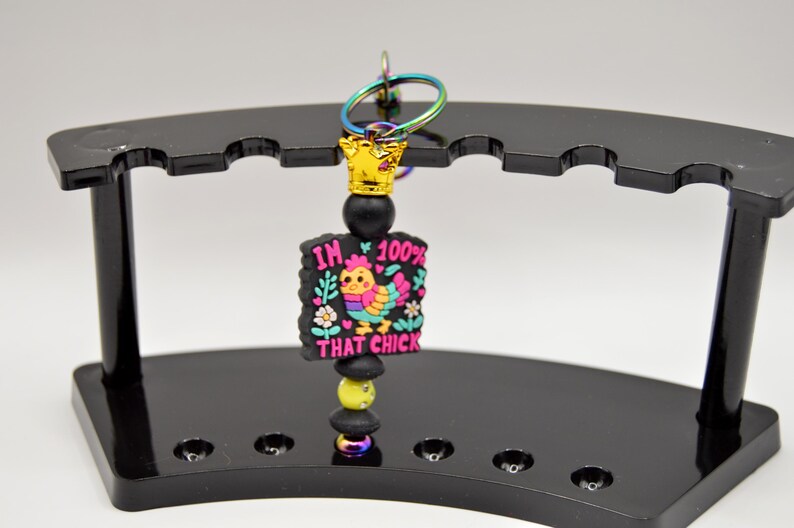May include: A black beaded keychain with a gold crown charm and a pink, yellow, and blue charm with the text "I'm 100% That Chick".