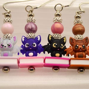 Könnte beinhalten: Handgefertigte Ohrringe mit Fledermausfiguren auf Miniatur-Buchanhängern. Jedes Paar zeigt ein einzigartiges Farbschema, darunter braune, violette, schwarze und graue Fledermäuse, mit glitzernden Perlenakzenten.