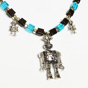 May include: A silver robot charm necklace with black and blue square beads. The necklace has three robot charms, one larger than the other two.