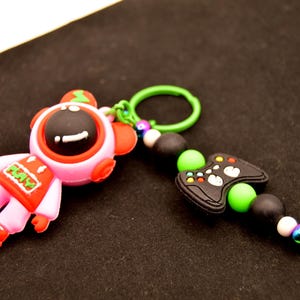 May include: A pink and red astronaut character keychain with a green and black video game controller charm. The astronaut has a black face with a white smile and the words "PLAY" on its chest. The keychain is attached to a green ring.