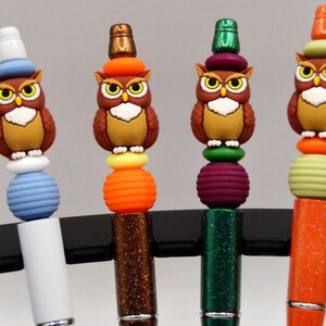 May include: A collection of decorative pens featuring owl-shaped toppers. Each pen has a unique color scheme, including brown, yellow, orange, green, and purple, with matching beads and glittery barrels. The owl figures have detailed eyes and hats.