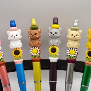 May include: Six colorful pens with cat-shaped toppers. Each pen features a different colored barrel and a cat figurine atop a sunflower design. The pens are arranged in a row, showcasing a variety of colors and designs.