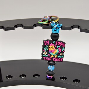 May include: A colorful keychain with a black and white charm that says "I'm 100% That Chick" with a cartoon chicken in the center. The charm is attached to a black beaded chain with a blue crown at the top.