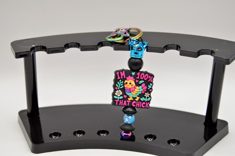 May include: A colorful keychain with a black and white charm that says "I'm 100% That Chick" with a cartoon chicken in the center. The charm is attached to a black beaded chain with a blue crown at the top.