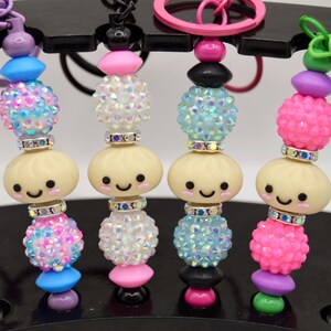May include: Four beaded keychains featuring a central cream-colored dumpling charm with a smiling face, flanked by colorful textured bubble beads, sparkling rhinestone spacers, and solid-colored accent beads attached to metal key rings.