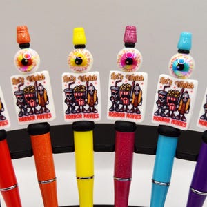 May include: A collection of colorful pens with decorative toppers. Each pen features a different color barrel and a unique topper with an eyeball design and a rectangular charm that reads "Let's Watch HORROR MOVIES".