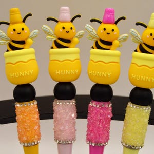 Puede incluir: Cuatro bolígrafos decorativos con abejas amarillas sobre botes de miel amarillos con la palabra "HUNNY". Los bolígrafos tienen cuentas negras y barriles de colores con joyas en naranja, rosa y amarillo.