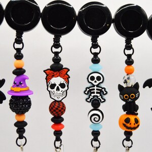 May include: A collection of black retractable badge holders with Halloween-themed charms. Charms include a spider, witch hat, skull, skeleton, black cat, pumpkin, bat, and vampire. Each charm is attached with beads.