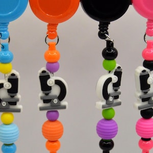 May include: A collection of colorful retractable badge holders. Each holder features a microscope charm and a beaded chain in various colors, including yellow, blue, orange, black, and pink. The charms are white, black, and gray.