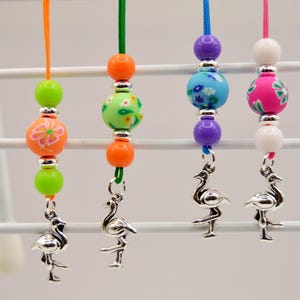 May include: Four beaded flamingo charms hang from colorful cords. The beads are green, orange, blue, purple, pink, and white, with floral and patterned designs. Each charm has a silver flamingo pendant.