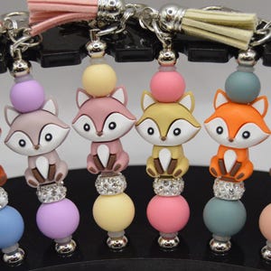 Puede incluir: Llaveros con dijes de zorro en varios colores, incluyendo marrón, naranja y rosa. Cada llavero tiene un diseño de cuentas, una borla y un cierre plateado. Los dijes de zorro tienen caras blancas y están sobre un diseño de cuentas.