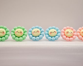 Può includere: Sei pomelli decorativi a forma di fiore. Ogni pomello presenta un volto sorridente al centro, circondato da piccole perline rotonde. I colori includono verde, azzurro e rosa.