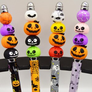 May include: Four pens featuring Halloween-themed bead toppers. Each pen has a unique barrel pattern and a stack of four beads, including jack-o'-lanterns, ghosts, bats, and spiders in orange, purple, black, green, yellow, and white colours.