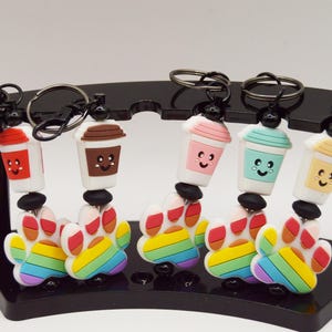 Op de afbeelding: Zes kleurrijke sleutelhangers met regenboog-pootafdrukcharms en cartoon-drankcharms. De charms zijn bevestigd aan zilveren sleutelringen en worden getoond op een zwarte standaard.
