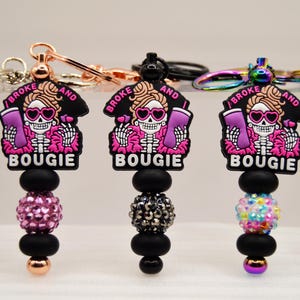 Puede incluir: Varios llaveros con un diseño de calavera con gafas de sol y el texto "I BROKE AND BOUGIE". Cada llavero tiene un diseño de cuentas de diferentes colores, incluyendo plata, rosa y oro.