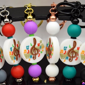 Puede incluir: Llaveros con colgantes de notas musicales. Cada llavero presenta un colgante redondo y blanco con una clave de sol y notas musicales coloridas. Los colgantes se combinan con cuentas de colores en rojo, morado, turquesa, blanco, gris y negro.
