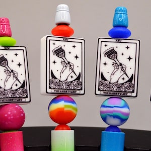 May include: A collection of colorful, decorative hair styling tools. Each tool features a small, rectangular card with a black and white illustration of a hand holding a brush, with the text "THE HAIRSTYLIST". The tools have various colored beads and toppers.