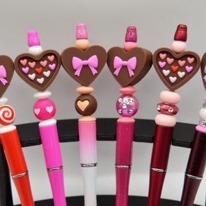 May include: A selection of decorative pens featuring heart-shaped toppers. The toppers are brown with pink bows and heart details. The pens are in various colors, including red, pink, and white, and are displayed on a black stand.
