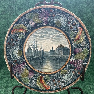 May include: Decorative plate with a central harbor scene, surrounded by a border of flowers, grapes, and fruit. The plate is displayed on a dark metal stand, set against a green patterned background.