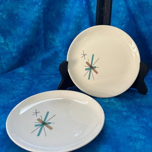May include: Two white ceramic plates with a mid-century modern design. Each plate features a teal, brown, and black starburst pattern. The plates are displayed on a blue background.