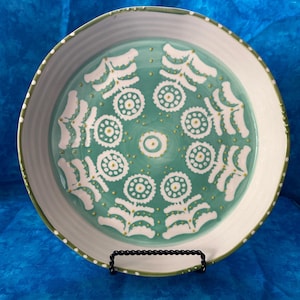 Puede incluir: Un plato de cer&aacute;mica decorativo con un centro turquesa con motivos florales blancos y un borde blanco. El plato tiene un borde verde y se exhibe sobre un soporte de metal negro. El dise&ntilde;o recuerda al arte popular.