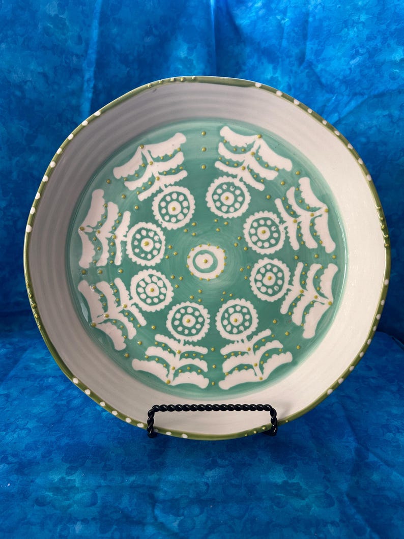 Puede incluir: Un plato de cer&aacute;mica decorativo con un centro turquesa con motivos florales blancos y un borde blanco. El plato tiene un borde verde y se exhibe sobre un soporte de metal negro. El dise&ntilde;o recuerda al arte popular.