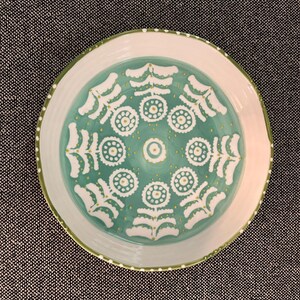 Puede incluir: Un plato redondo de cer&aacute;mica con borde blanco y centro turquesa. El centro presenta un patr&oacute;n floral blanco con detalles amarillos. El borde tiene un borde verde y peque&ntilde;os puntos blancos. Este plato decorativo es adecuado para servir alimentos.