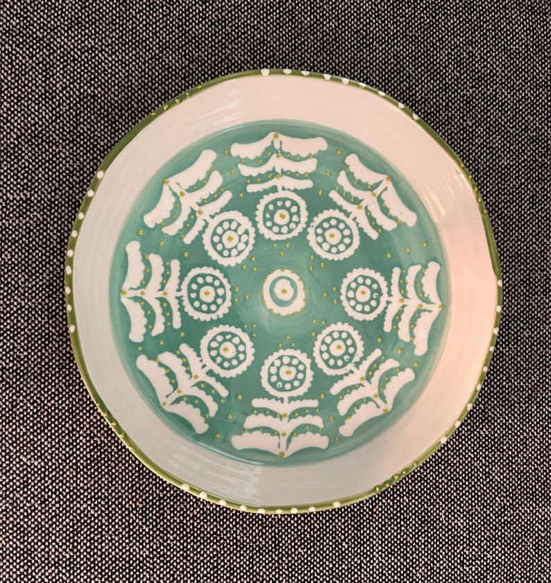 Puede incluir: Un plato redondo de cer&aacute;mica con borde blanco y centro turquesa. El centro presenta un patr&oacute;n floral blanco con detalles amarillos. El borde tiene un borde verde y peque&ntilde;os puntos blancos. Este plato decorativo es adecuado para servir alimentos.
