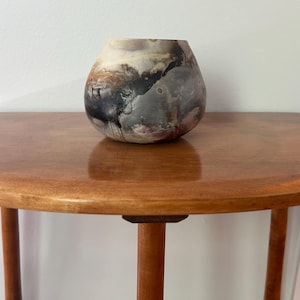 May include: A handcrafted, organic-shaped ceramic vase with a unique, abstract design. The vase features a blend of colors, including black, gray, brown, and cream, creating a textured, artistic appearance. The vase is displayed on a wooden table.