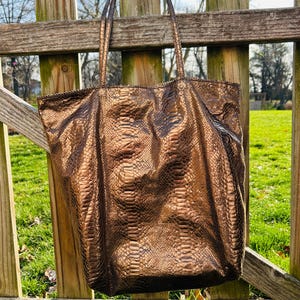 May include: A large, brown tote bag with a snakeskin pattern hangs on a wooden fence. The bag has long straps and a spacious interior. The bag's color is a metallic bronze, and the texture is visible.