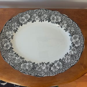 May include: An oval ceramic serving platter with a white center and a wide, scalloped border featuring a detailed black and white floral pattern of roses and leaves against a stippled background, creating a high-contrast decorative design.