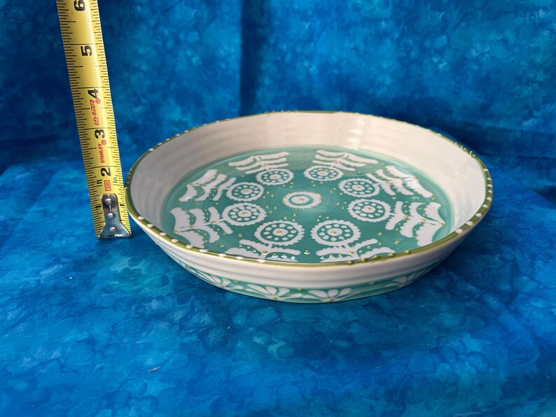Puede incluir: Un plato redondo de cer&aacute;mica con borde blanco e interior turquesa decorado con motivos florales blancos. El plato mide aproximadamente 6,3 cm de alto y 20 cm de di&aacute;metro. Una cinta m&eacute;trica amarilla est&aacute; al lado del plato.