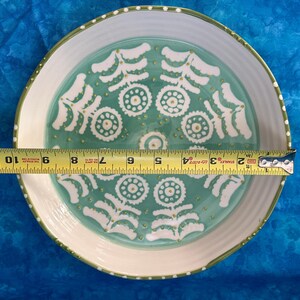 Puede incluir: Un plato decorativo de cer&aacute;mica con borde blanco y centro turquesa. El plato presenta un patr&oacute;n repetitivo de dise&ntilde;os florales blancos y detalles dorados. El plato mide aproximadamente 25 cm de di&aacute;metro.
