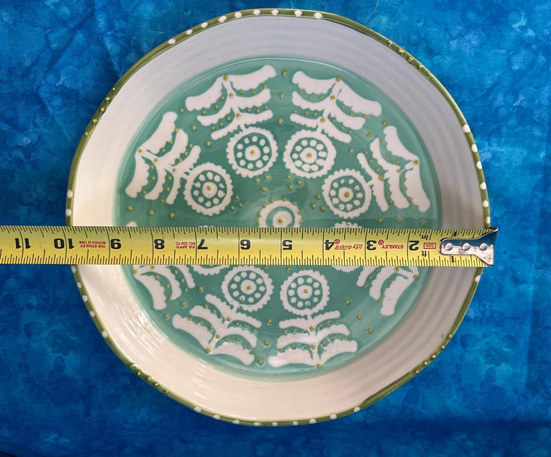 Puede incluir: Un plato decorativo de cer&aacute;mica con borde blanco y centro turquesa. El plato presenta un patr&oacute;n repetitivo de dise&ntilde;os florales blancos y detalles dorados. El plato mide aproximadamente 25 cm de di&aacute;metro.