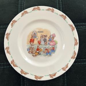 May include: White ceramic plate with a whimsical illustration of rabbits in a train station scene. The plate features a border of brown rabbits and a green dotted line. The center scene includes the word "TICKETS".