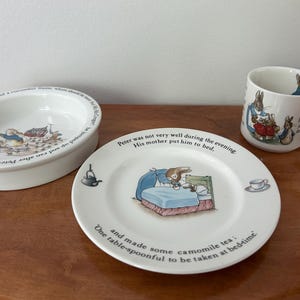 May include: A set of three ceramic pieces featuring illustrations of Peter Rabbit. The set includes a white oval dish, a white plate with text, and a white mug. The plate shows Peter Rabbit in bed. The mug has a handle and a scene of Peter Rabbit with other rabbits.