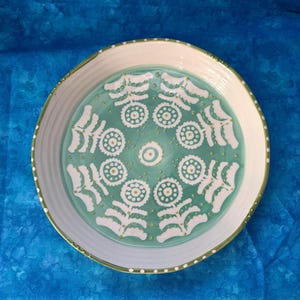 Puede incluir: Una fuente redonda de cer&aacute;mica con borde blanco y centro turquesa. El centro presenta un patr&oacute;n floral blanco con detalles dorados. El borde tiene un borde verde con puntos blancos.