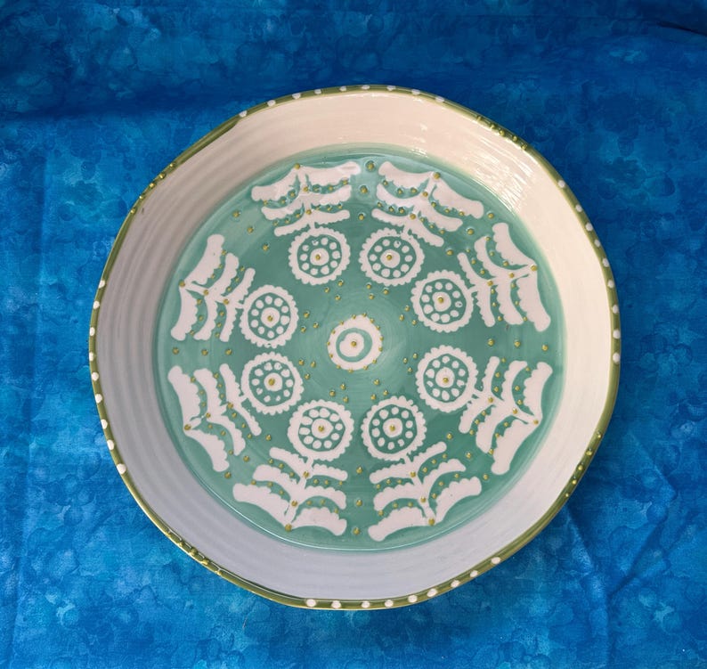Puede incluir: Una fuente redonda de cer&aacute;mica con borde blanco y centro turquesa. El centro presenta un patr&oacute;n floral blanco con detalles dorados. El borde tiene un borde verde con puntos blancos.