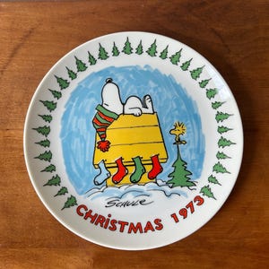 May include: A white ceramic decorative plate featuring a cartoon illustration of Snoopy lying on a yellow doghouse with a striped hat, Woodstock, and four hanging stockings. The border has green pine trees, and the text reads Christmas 1973.