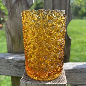 May include: A vintage amber-colored glass votive candle holder. The glass has a textured, starburst pattern. The candle holder is cylindrical with a scalloped top edge. It sits on a wooden post outdoors.