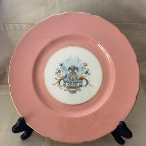 May include: A pink ceramic plate with a scalloped edge and gold trim. The center features a detailed illustration of a fountain with birds and flowers. A decorative piece for serving or display.
