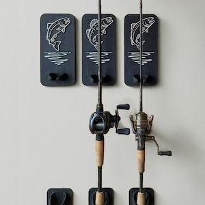 May include: A set of three black fishing rod holders with white fish illustrations and wave designs. Two fishing rods with reels are mounted on the holders. The holders are mounted on a light gray wall.