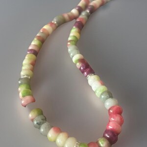Puede incluir: Un collar de cuentas de colores con una variedad de cuentas redondas en tonos rosa, verde, blanco y morado. Las cuentas est&aacute;n ensartadas con un espaciador plateado. El collar se muestra sobre un fondo gris.
