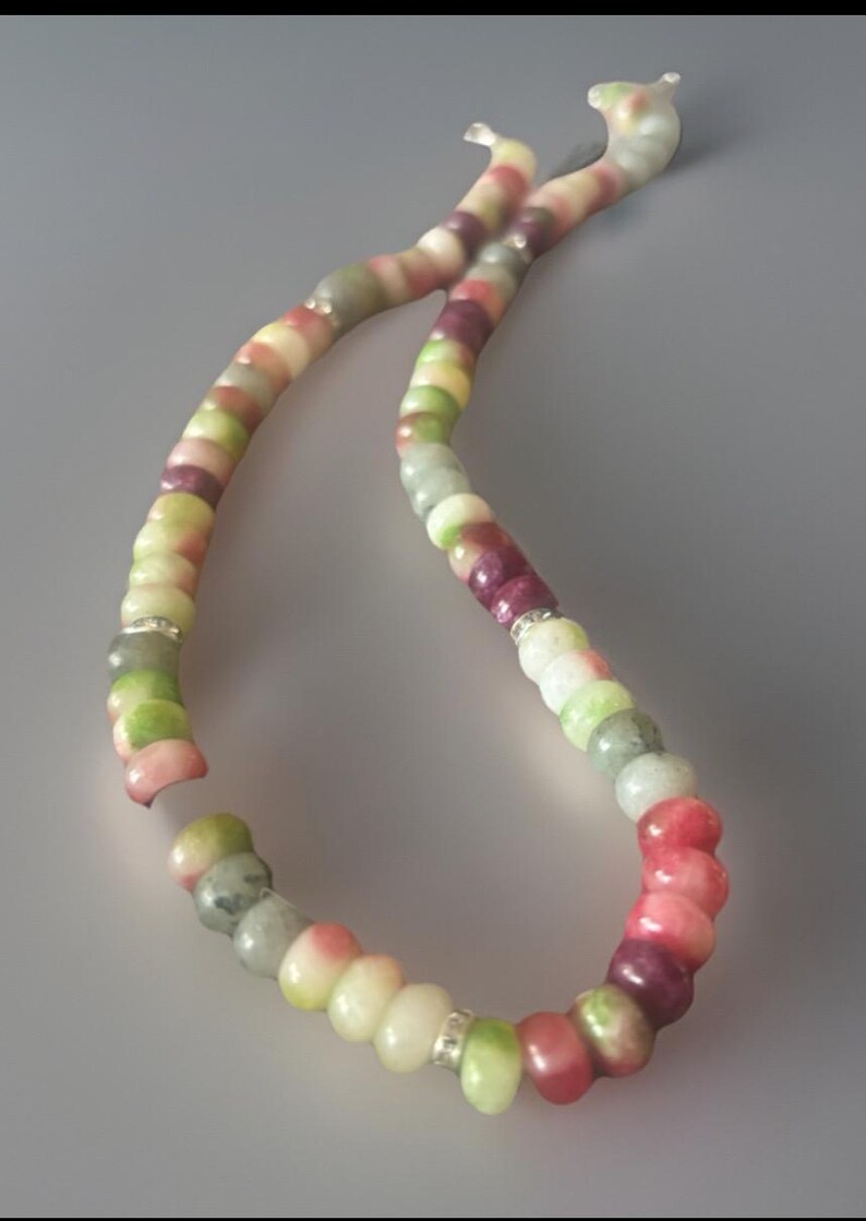 Puede incluir: Un collar de cuentas de colores con una variedad de cuentas redondas en tonos rosa, verde, blanco y morado. Las cuentas est&aacute;n ensartadas con un espaciador plateado. El collar se muestra sobre un fondo gris.
