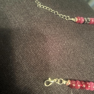 Peut inclure: Un collier avec une cha&icirc;ne et un fermoir argent&eacute;s. Le collier pr&eacute;sente une s&eacute;rie de perles roses et grises. La cha&icirc;ne est reli&eacute;e aux perles par du fil d'argent. Le collier est sur une surface gris fonc&eacute;.