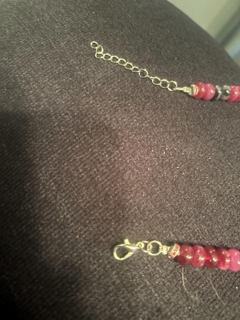 Peut inclure: Un collier avec une cha&icirc;ne et un fermoir argent&eacute;s. Le collier pr&eacute;sente une s&eacute;rie de perles roses et grises. La cha&icirc;ne est reli&eacute;e aux perles par du fil d'argent. Le collier est sur une surface gris fonc&eacute;.