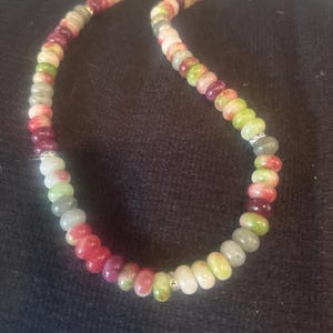 Puede incluir: Un collar de cuentas de colores con cuentas de piedras preciosas en tonos rojos, verdes, blancos y grises. El collar tiene un cierre dorado y peque&ntilde;os detalles plateados. Las cuentas tienen aproximadamente 0,8 cm de di&aacute;metro.