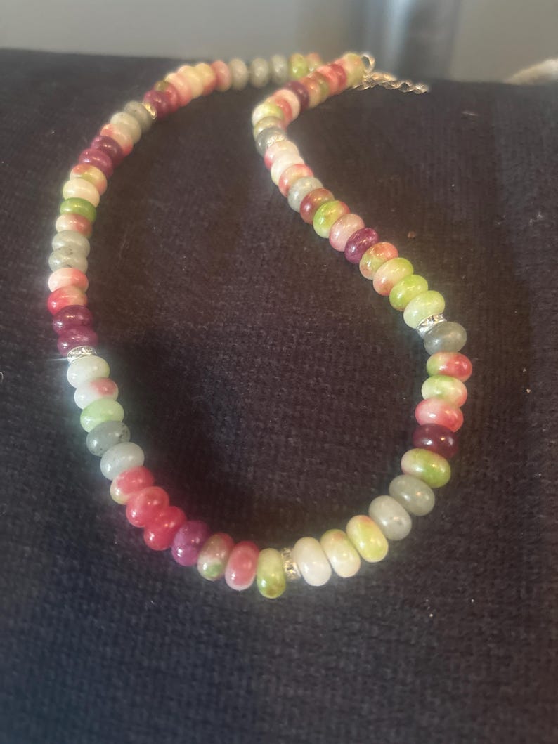Puede incluir: Un collar de cuentas de colores con cuentas de piedras preciosas en tonos rojos, verdes, blancos y grises. El collar tiene un cierre dorado y peque&ntilde;os detalles plateados. Las cuentas tienen aproximadamente 0,8 cm de di&aacute;metro.