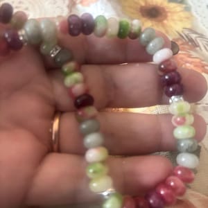 Puede incluir: Una pulsera de cuentas coloridas con cuentas de piedras preciosas en tonos rojos, rosas, verdes y grises. La pulsera est&aacute; ensartada en un cord&oacute;n flexible e incluye detalles plateados. Las cuentas miden aproximadamente 0,6 cm de di&aacute;metro.